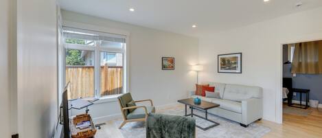 A modern living room with a flat-screen TV, a white sofa, a coffee table, and a large window with blinds.