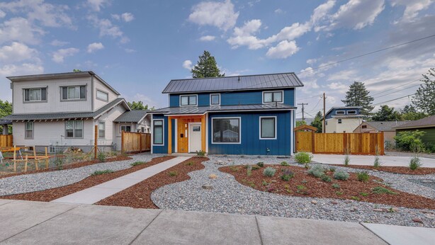 Une maison à deux étages, à l’extérieur bleu, avec une cour avant comprenant un chemin de gravier et quelques petites plantes.