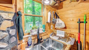 A rustic wooden cabin interior with a stone wall, wooden shelves with potted plants, a sink, and a window with a view of greenery.
