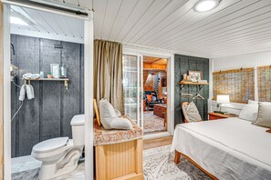 A hotel room with a wooden floor, a bed, a nightstand, a chair, a desk, a lamp, a shelf with towels, a framed picture, and a sliding door to a living area. - Cozy Nature Escape! Roaring Gap Cabin w/ Patio (Roaring Gap)