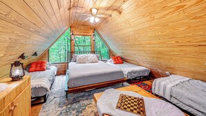 A cozy attic bedroom with wooden walls, a sloped ceiling, and a large window offering a view of the forest. There are two beds, a wooden dresser, and a chessboard on the floor.