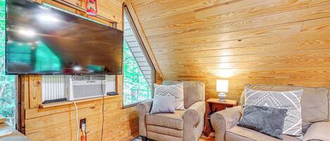 L’intérieur d’un chalet accueillant, doté d’un grand téléviseur, de deux fauteuils, d’un plafond en bois et d’une fenêtre donnant sur un paysage verdoyant.