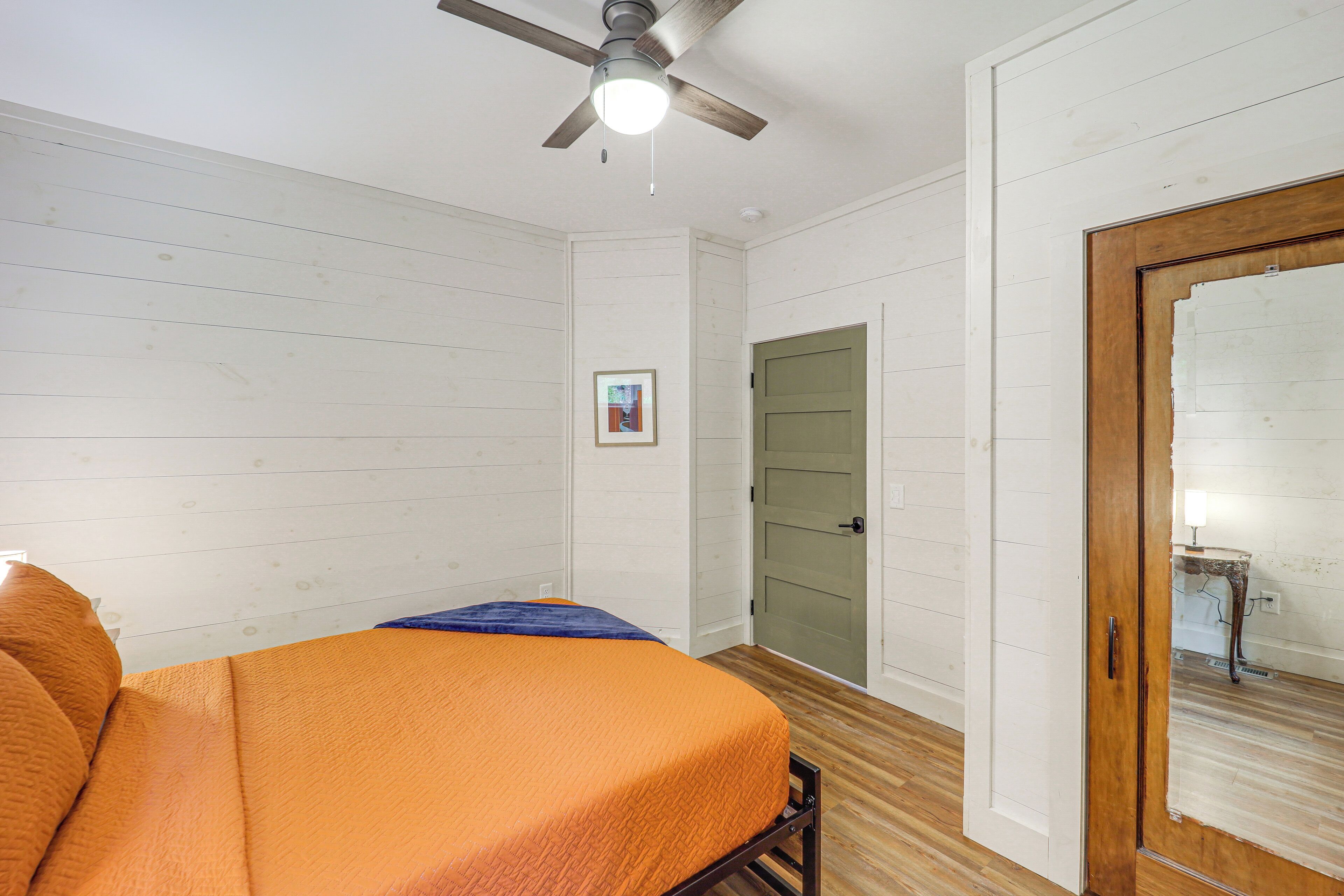Une pièce aux murs recouverts de panneaux blancs, avec un ventilateur de plafond, un lit avec une couvre-lit orange, un tableau encadré accroché au mur et une porte verte.