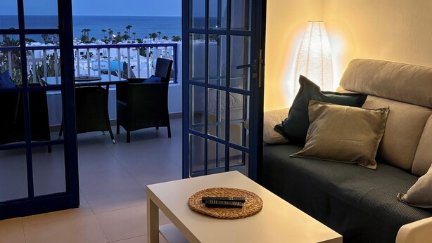 A balcony with a view of the sea, a sofa with cushions, and a table with a remote control.