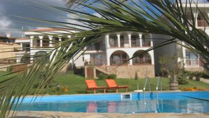 Una piscina con lettini, una palma e un edificio sullo sfondo con finestre ad arco.
