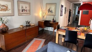 A modern living room with a wooden coffee table, a sofa, and a dining area with a wooden table and chairs. There are framed artworks on the walls and a red pendant light above the dining table.