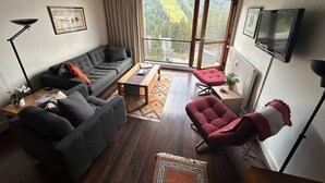 A modern living room with a grey sofa, red armchair, wooden coffee table, and a large window offering a view of a forested area.