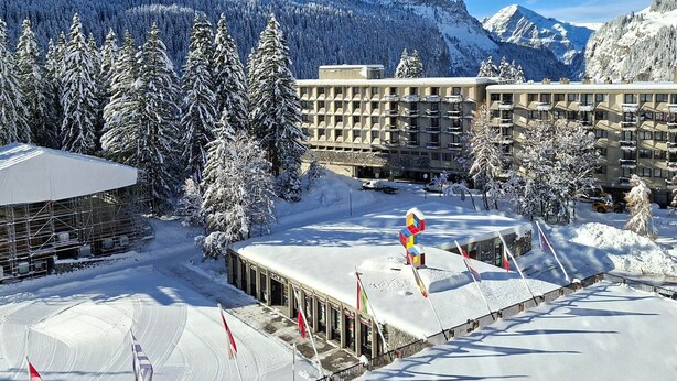 A snow-covered hotel with a mountain backdrop, featuring a modern building with multiple floors and a flat roof.