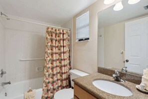 A bathroom with a shower curtain, a toilet, a sink with a mirror, and a window with blinds. - A-108 - Resort View and Corner Unit (Naples)