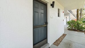A white exterior wall with a dark grey door, a small window, and a number '108' sign. - A-108 - Resort View and Corner Unit (Naples)