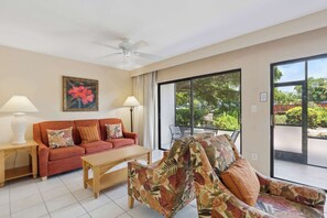 A living room with a sofa, a coffee table, and a ceiling fan. There is a large window with a view of greenery outside. - A-108 - Resort View and Corner Unit (Naples)