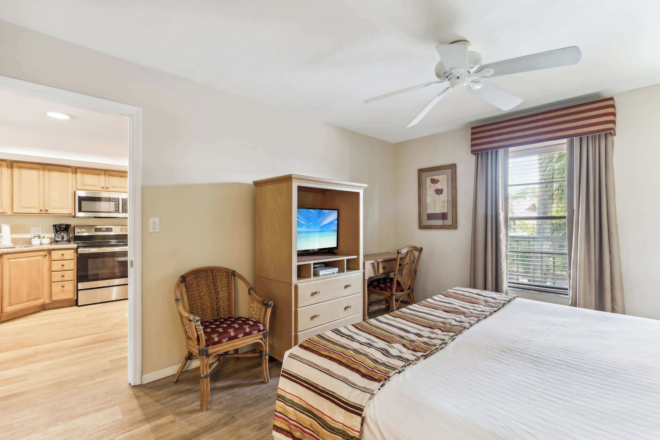 Habitación de hotel con cama, televisor, escritorio y silla. Se observa una kitchenette al fondo.