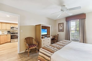 A hotel room with a bed, a TV, a desk, and a chair. There is a kitchenette in the background.