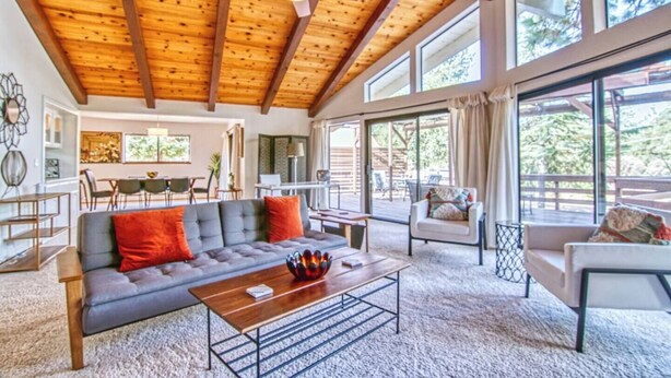 Un salon spacieux avec un plafond haut, un plafond en bois, un canapé gris agrémenté de coussins rouges, une table basse en bois et une grande fenêtre offrant une vue sur l’extérieur.