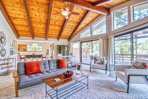 Un salon spacieux avec un plafond haut, un plafond en bois, un canapé gris agrémenté de coussins rouges, une table basse en bois et une grande fenêtre offrant une vue sur l’extérieur.