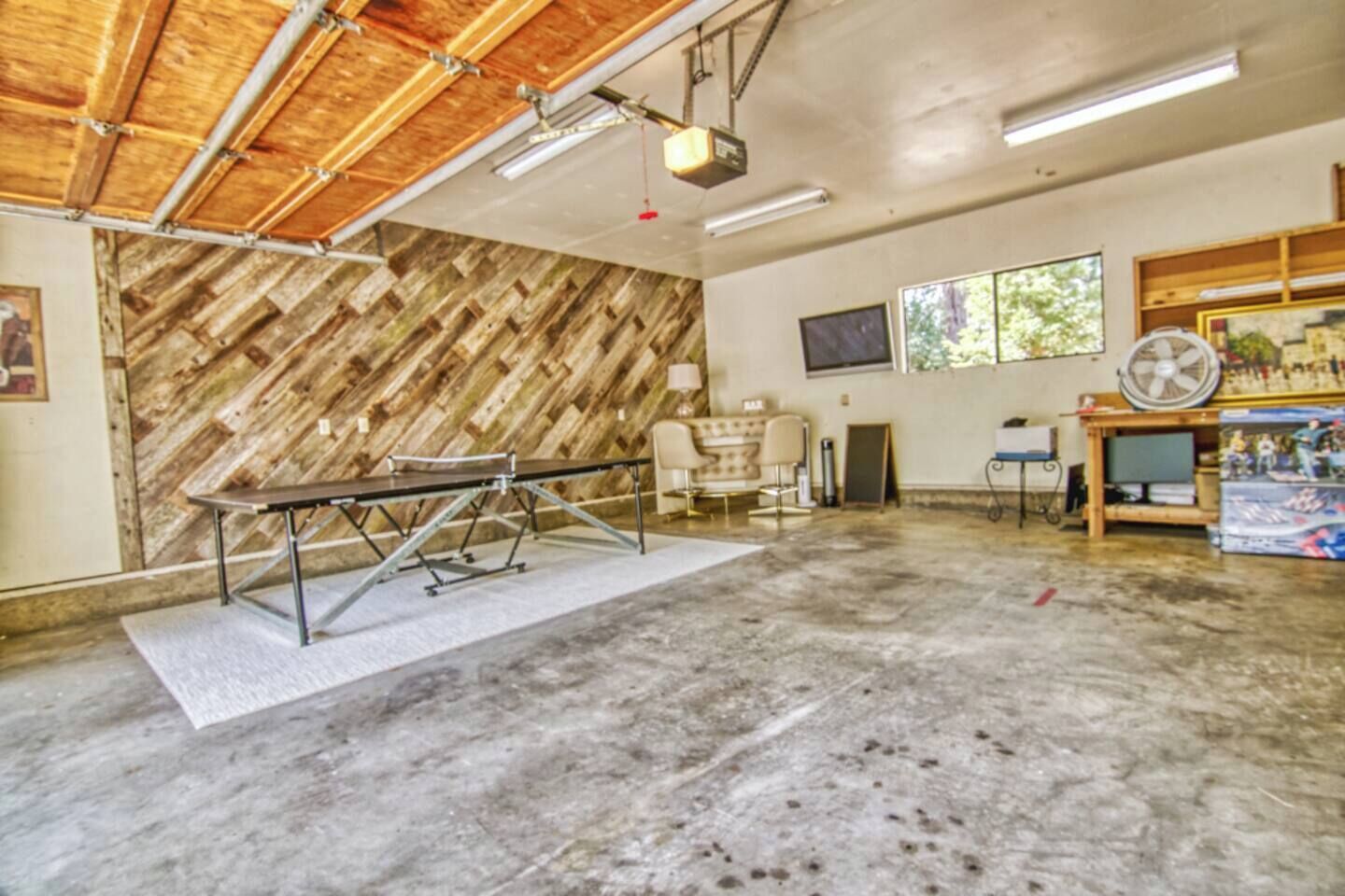 Une pièce spacieuse avec un sol en béton, un mur recouvert de lambris en bois, une table avec des chaises, un coin bar et une grande fenêtre.