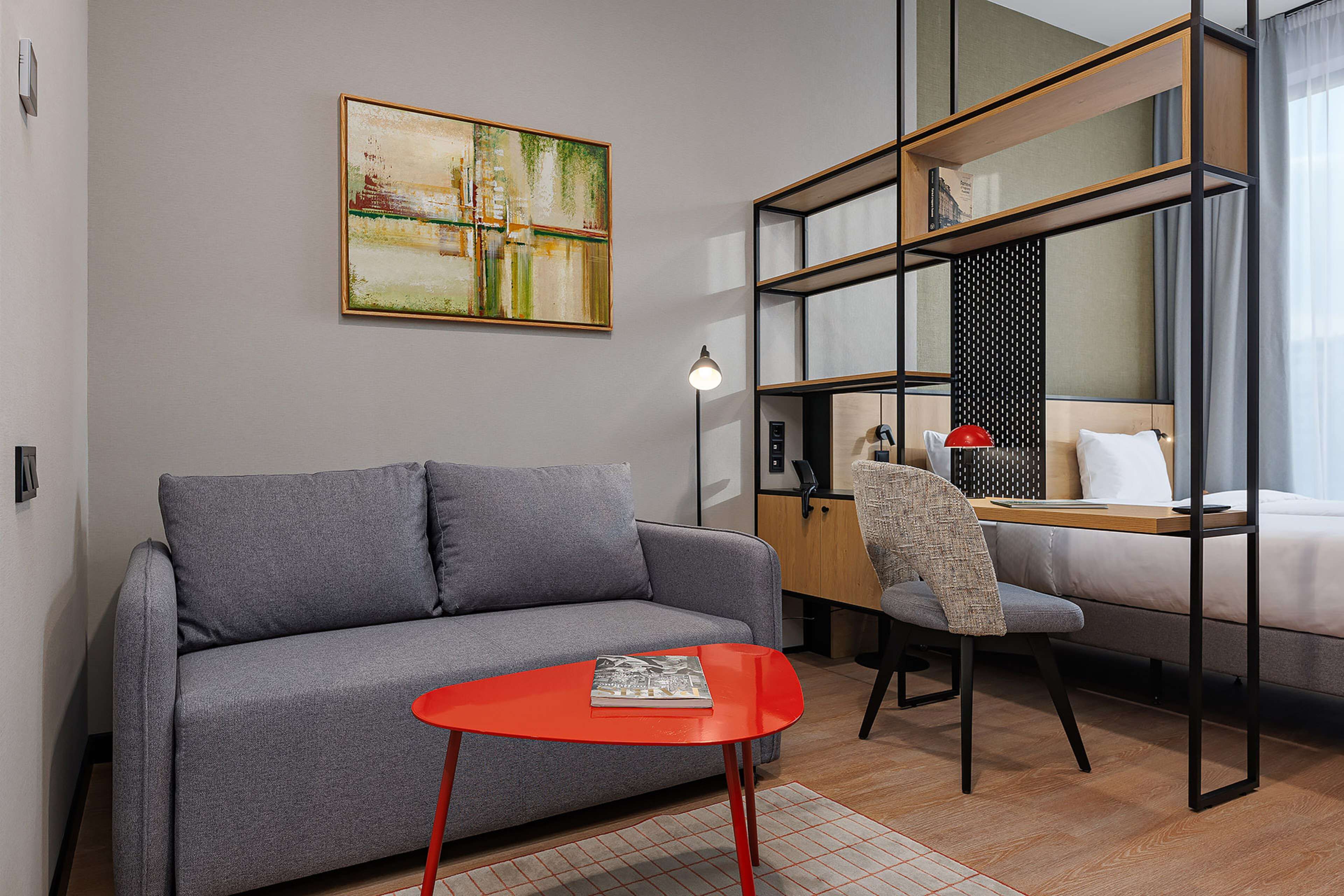 Una habitación de hotel moderna con un sofá gris, una mesa roja, una estantería, una cama y un cuadro en la pared.