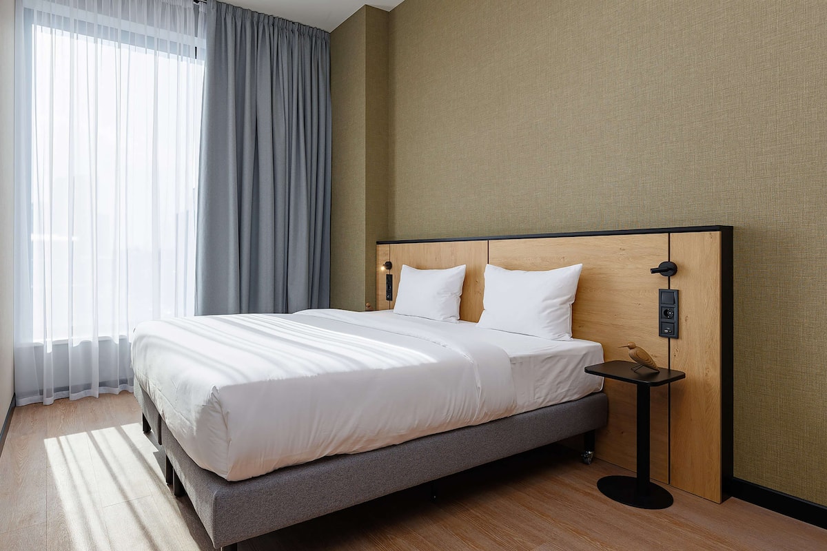 Habitación de hotel con una cama grande, una mesita de noche y vistas al exterior a través de cortinas traslúcidas.