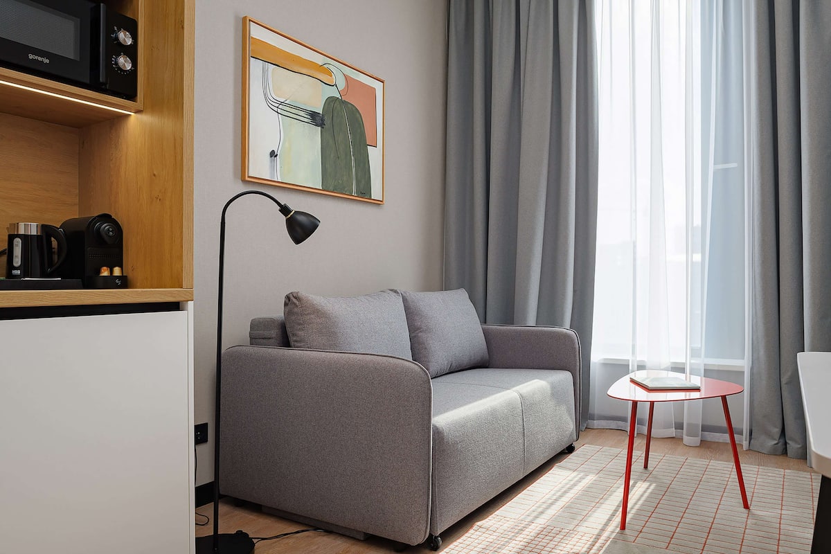 Una sala moderna con un sofá gris, una mesita roja pequeña, una lámpara de pie negra y una zona de cocina con microondas y cafetera.