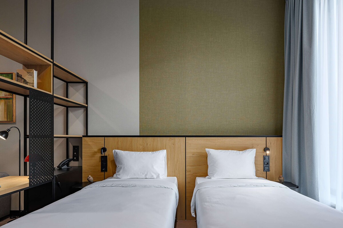 A hotel room with two beds, a desk, a bookshelf, and a large window with curtains.