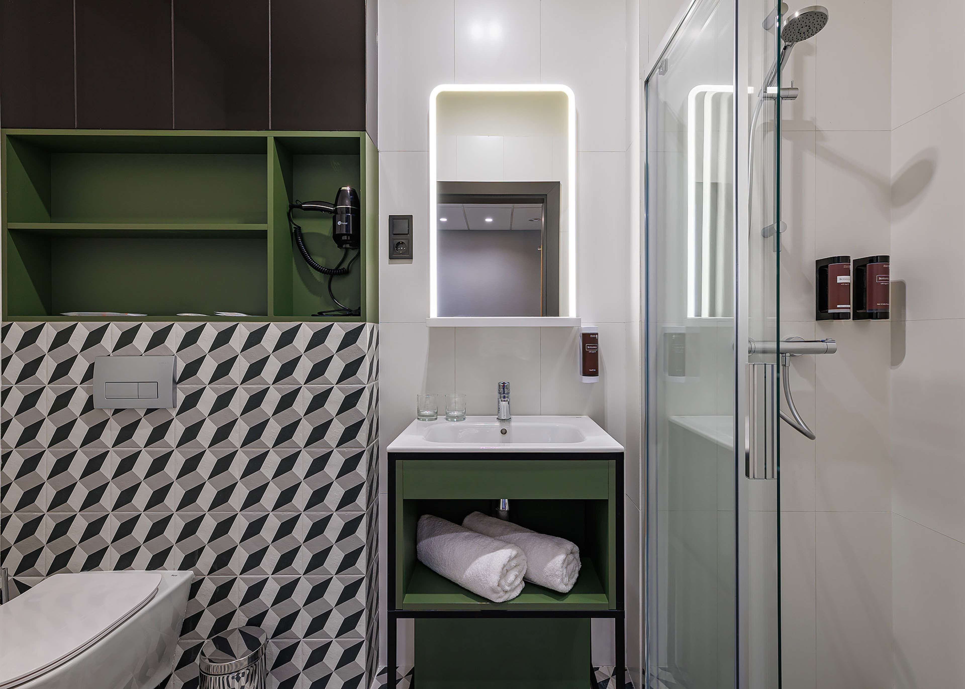 A modern bathroom with a geometric patterned wall, a glass shower enclosure, a green vanity with a mirror, and a toilet.