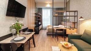 A modern hotel room with a dining area, a bed, a TV, a bookshelf, and a small table with fruit.