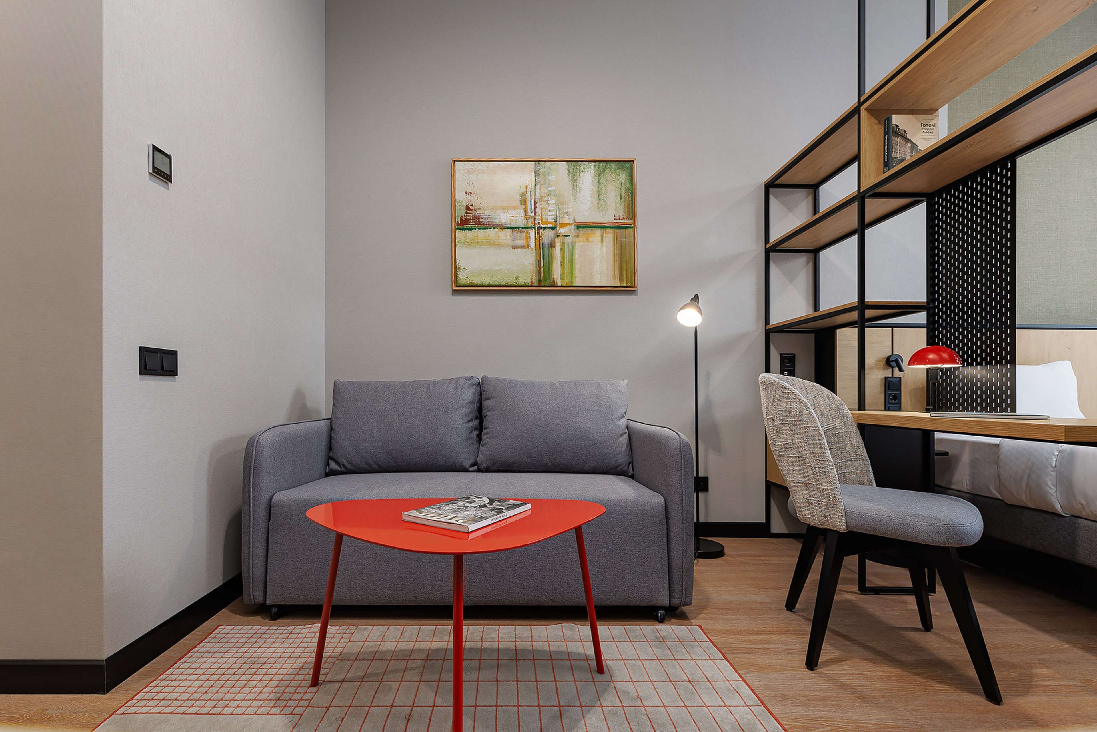 A modern living room with a grey sofa, a red table, a patterned armchair, and a bookshelf with a red lamp.