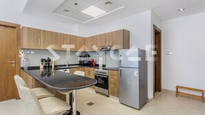 A modern kitchen with wooden cabinets, a stainless steel refrigerator, an oven, and a dining area with two chairs. - Staycae- National Bonds Residences  (Dubai)