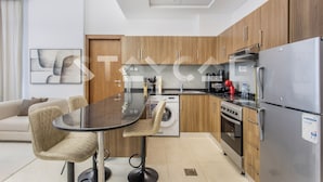 A modern kitchen with wooden cabinets, a stainless steel refrigerator, a built-in oven, and a washing machine. There is a dining table with chairs and a sofa in the adjacent living area. - Staycae- National Bonds Residences  (Dubai)
