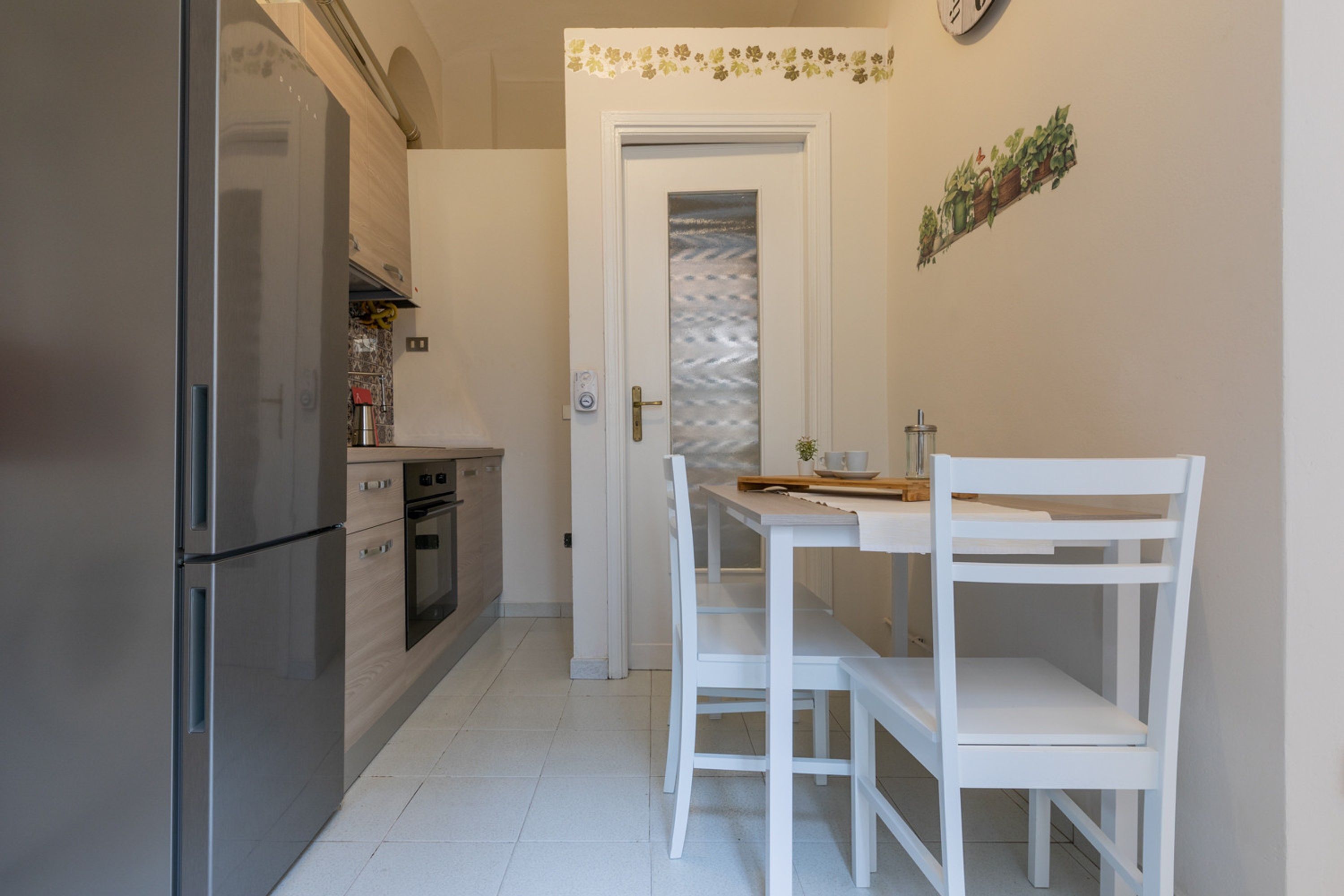 Cocina compacta con refrigerador, horno y muebles blancos. Una mesa de comedor con sillas se encuentra cerca de una puerta con un panel decorativo.