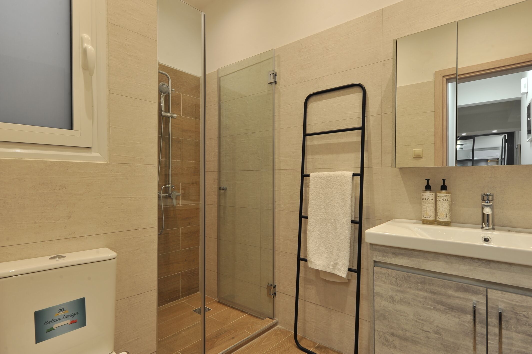 A modern bathroom with a glass-enclosed shower, a black towel rack, a sink with toiletries, and a mirror reflecting the room.
