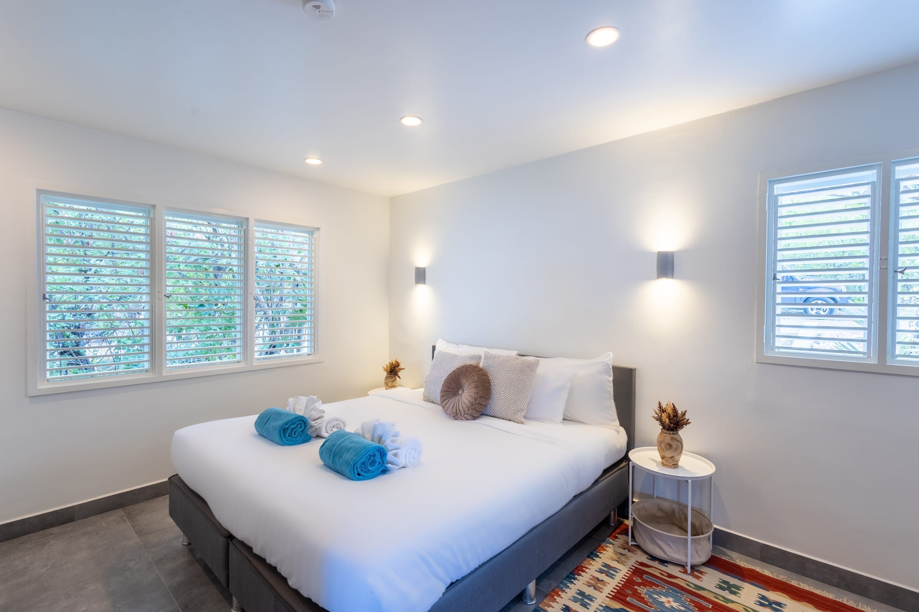 A modern hotel room with a large bed, two windows with blinds, a small bedside table with a vase, and a patterned rug.