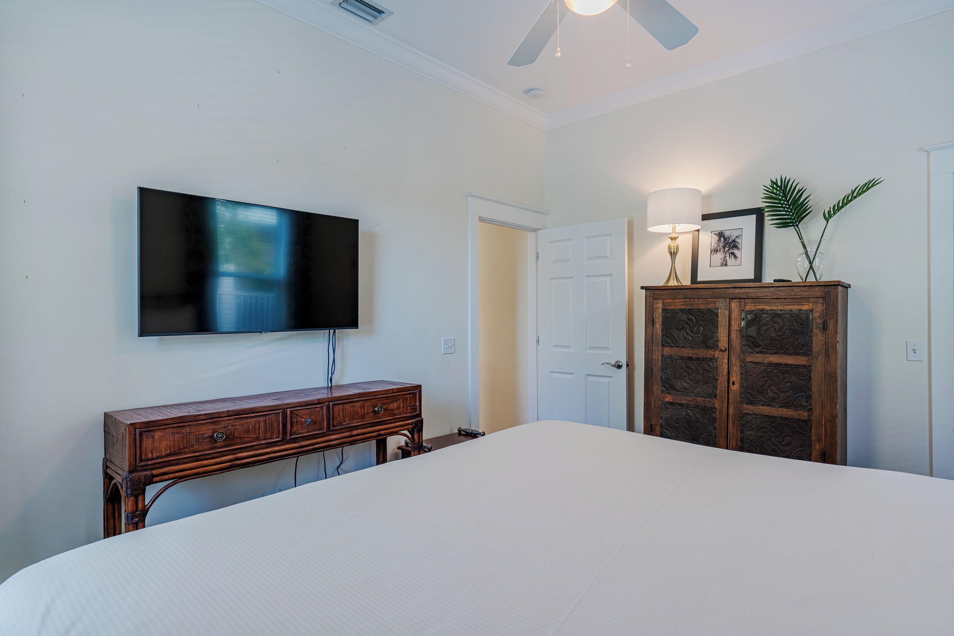 A bedroom with a bed, a wooden dresser, a television mounted on the wall, a door, and a ceiling fan.