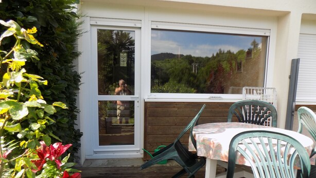 An outdoor patio with a wooden table and chairs, a sliding glass door, and a view of greenery and hills. - Location Saisonnière en Bord de Loire (Coubon)