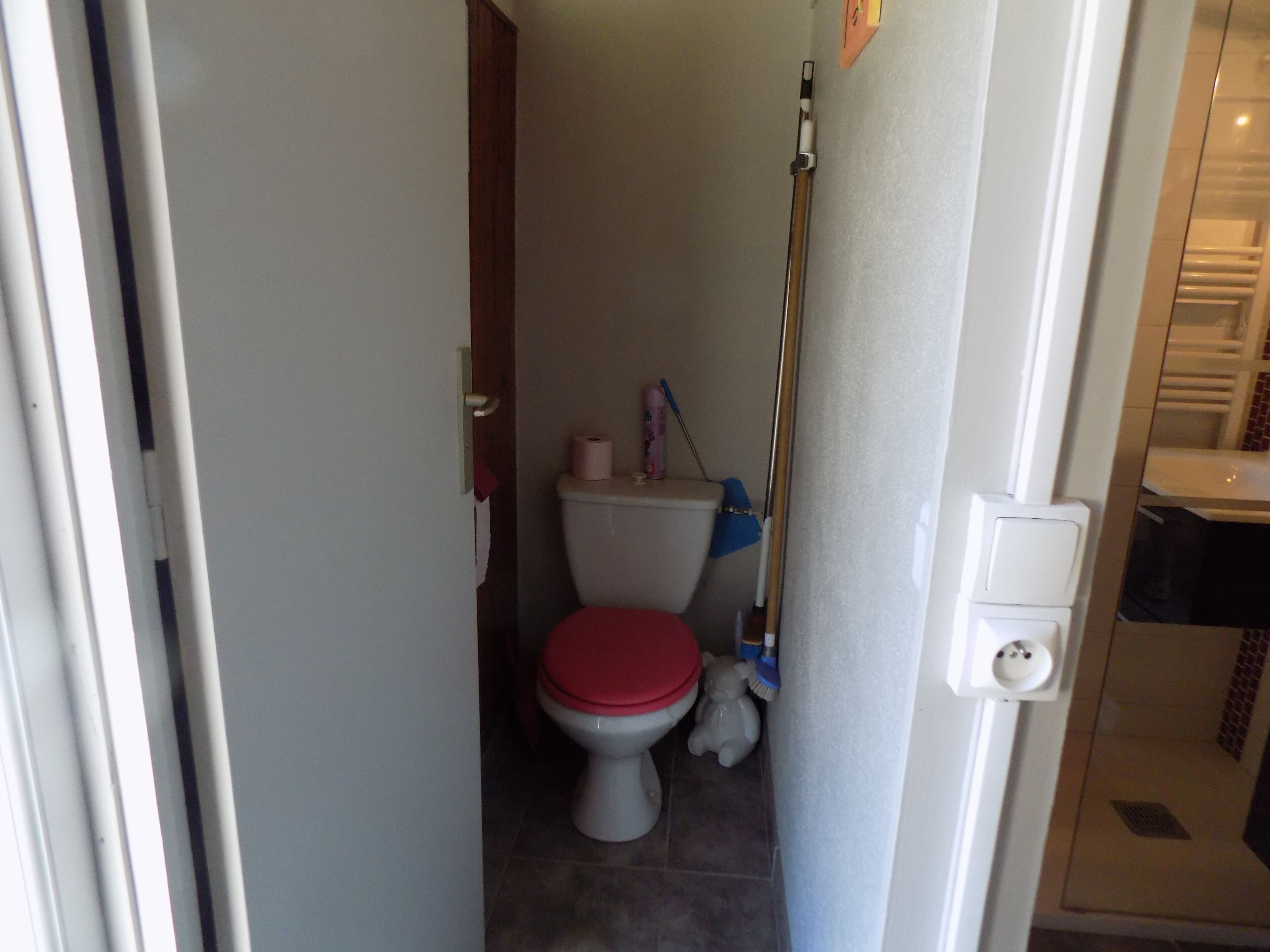 A small bathroom with a toilet, a red seat, a roll of toilet paper, and a mop leaning against the wall.