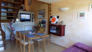 A cozy living room with a wooden dining table, chairs, a sofa, and a TV mounted on the wall. - Location Saisonnière en Bord de Loire (Coubon)