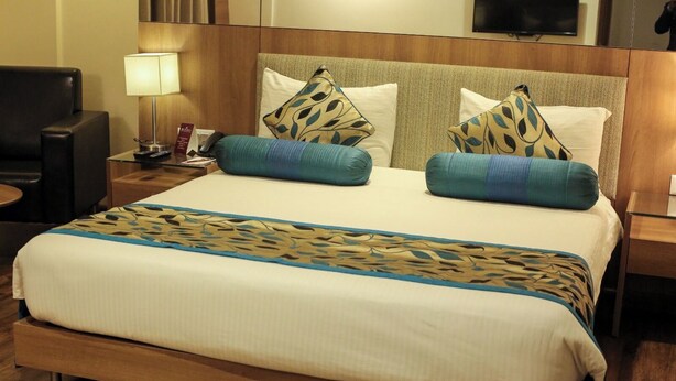 Habitación de hotel con una cama grande, dos almohadas con motivos de hojas, una mesita de noche con lámpara y un televisor en la pared.