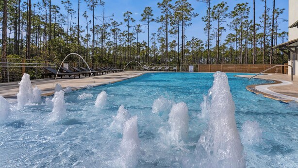 A swimming pool with water jets, surrounded by lounge chairs and a wooden deck, with tall pine trees in the background. - Pinewood30a 146 (Santa Rosa Beach)