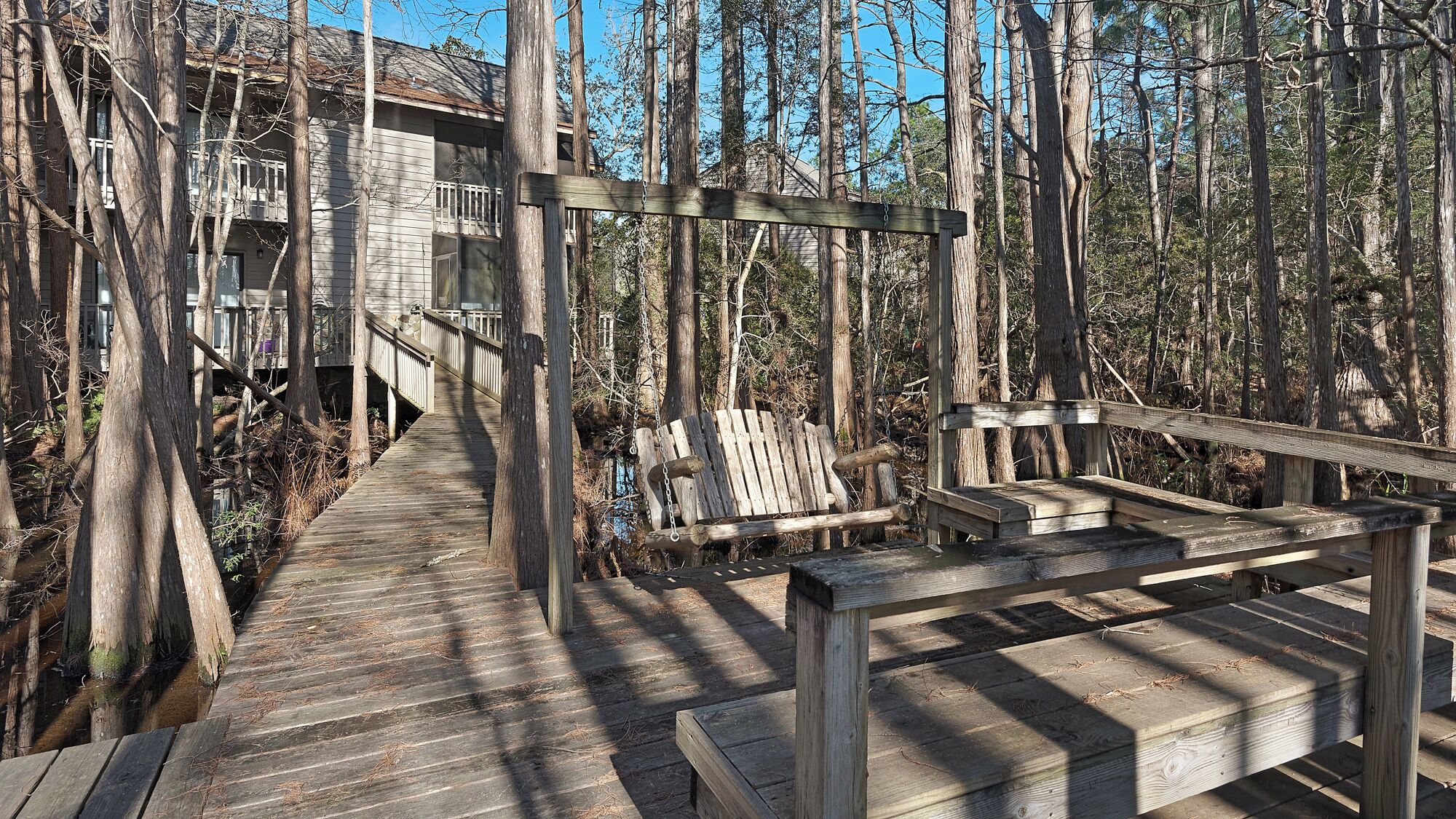 A wooden boardwalk leading to a building with a balcony, surrounded by trees.