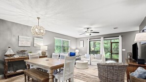 Un vaste espace de vie comprenant une table à manger en bois, des chaises blanches, un ventilateur de plafond et une grande fenêtre donnant sur un paysage verdoyant.