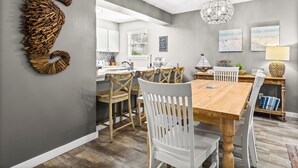 Un coin repas avec une table en bois et des chaises blanches, un lustre et une œuvre d’art murale représentant un hippocampe.