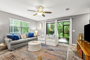 Un salon avec un canapé gris, deux fauteuils, une table basse, un ventilateur de plafond et une grande fenêtre avec des rideaux.