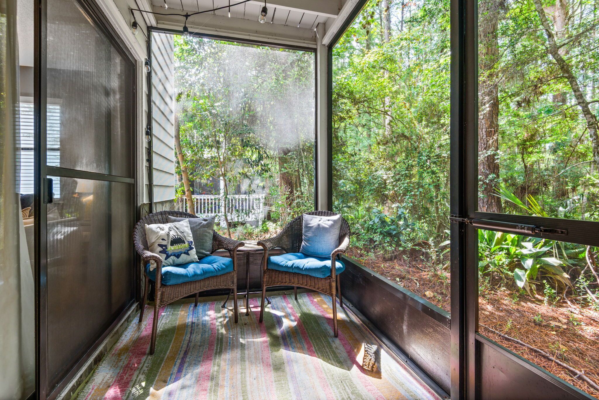 A screened porch with wicker furniture and a colorful rug, overlooking a lush garden.