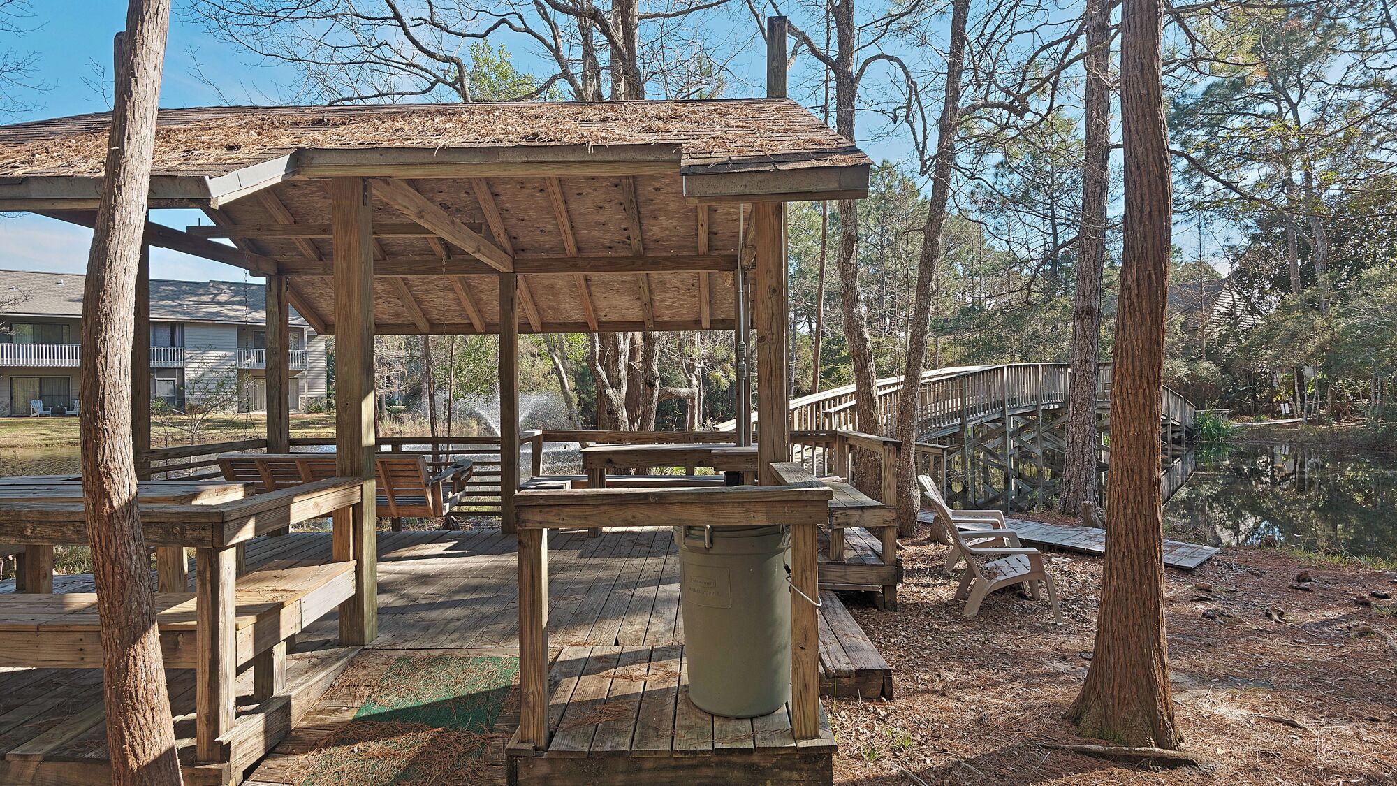 A wooden deck with picnic tables and a trash can, surrounded by trees and a wooden bridge over a pond.