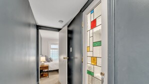 Ein Flur, der in ein Schlafzimmer mit Bett, Nachttisch, Lampe und Fenster führt.