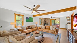 A spacious living room with a large sectional sofa, a glass coffee table, and a fireplace. There are two ceiling fans, wooden beams on the ceiling, and a television on a wooden cabinet. - The Getaway 4 Bedroom Home (Pittsfield)