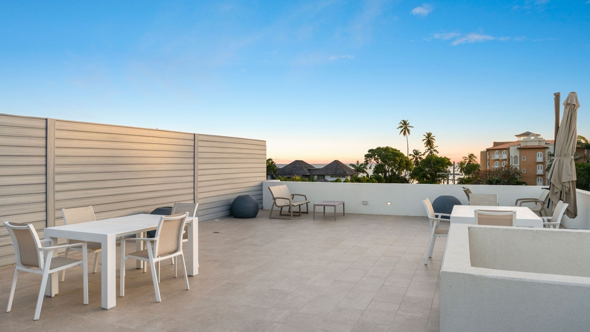 Un patio exterior con mobiliario blanco, un muro bajo y vistas a edificios y palmeras.