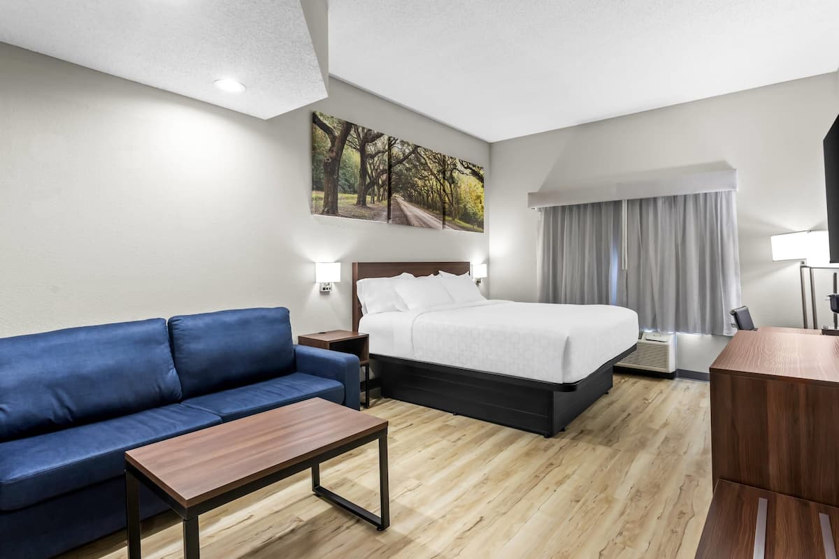 A hotel room with a bed, a blue sofa, a wooden coffee table, a painting on the wall, and a window with curtains.