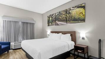 Habitación de hotel con una cama grande, una silla azul, una mesita de noche, una lámpara y un cuadro de un bosque en la pared.