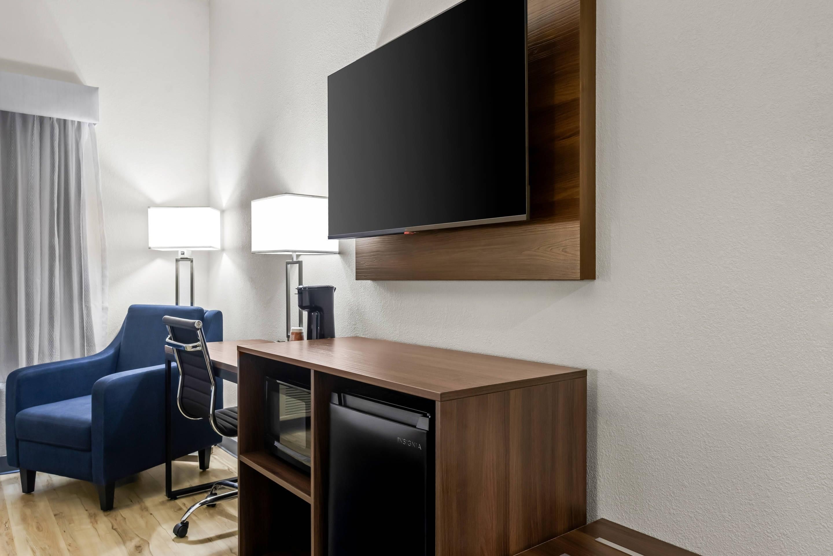 Habitación de hotel con televisión de pantalla plana en la pared, un escritorio con silla, cafetera y lavavajillas.